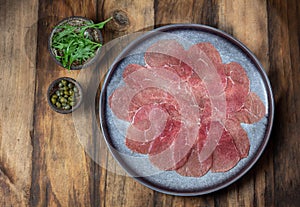 Marbled beef for carpaccio with ingredients arugula and capers on gray plate, wooden background, top view