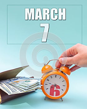 March 7th. Hand holding an orange alarm clock, a wallet with cash and a calendar date. Day 7 of month.