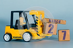march 27th. Day 27 of month, Construction or warehouse calendar. Yellow toy forklift load wood cubes with date. Work planning and