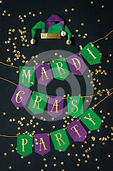 Mardi Gras party golden lettering on paper handmade green and purple festive garland, black background, confetti, jesters hat.