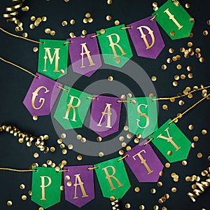 Mardi Gras party golden lettering on paper handmade green and purple festive garland, black background, confetti, jesters hat.