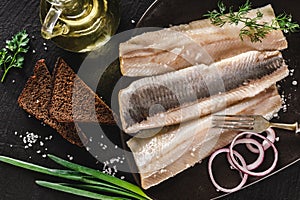 Marinated fillet mackerel or fillet herring fish with spices, greens and slice of bread on plate over dark stone background