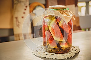 Marinated or pickled watermelon with herbs in a glass jar on a wooden table in a kitchen.