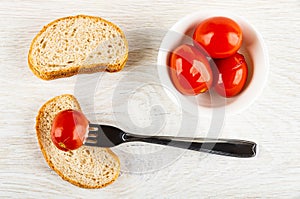 Marinated tomato strung on fork on bread, pieces of bread, bowl with tomatoes on table. Top view