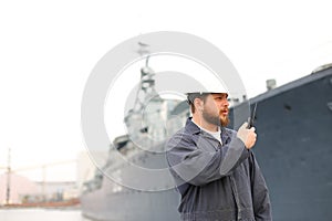 Marine captain talking by walkie talkie radio and standing near offshore vessel in bakground.