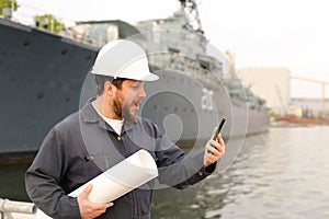 Marine engineer in helmet holding VHF walkie talkie and papers near vessel in background.