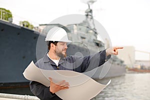 Maritime deckhand reading blueprints, showing by forefinger and standing near vessel.