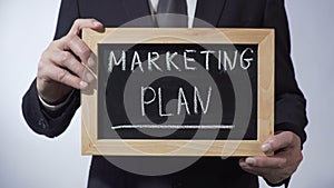 Marketing plan written on blackboard, man in black suit holding sign, business