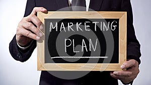 Marketing plan written on blackboard, man in black suit holding sign, business