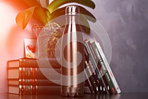 Maro of color thermo bottle over background of books and green flower. With sunlight effect