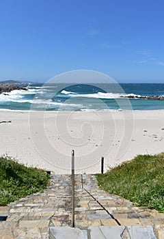 Beach with stone stairs, white sand and wild sea with waves, foam, and blue sky. A Marosa Beach, Burela, Lugo, Galicia, Spain.