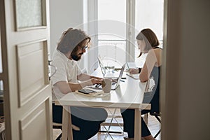 Married couple freelancers sit at table in living room with laptops and working from home office