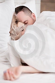 Married couple, man and woman, are lying in bed, hugging and sleeping on white bedding. Hiding their faces under blanket