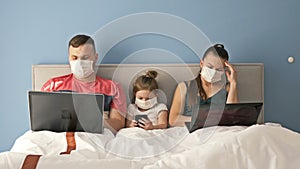 Married couple in medical masks lying on a bed with laptops. Little girl with a mobile phone in her hands. Pastime