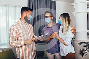 Married couple in medical masks standing in the new house with male real-estate agent. Pandemic concept
