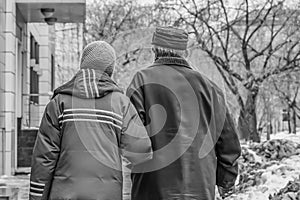 Married couple of pensioners walking arm-in-arm in the street with houses and trees and dirty snow in winter and an elderly woman