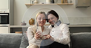 Married couple relax on sofa at home cuddle use smartphone