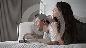 A married couple works remotely or makes online purchases on a laptop while lying on the couch at home
