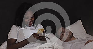 Married multi-ethnic couple lying in bed at night. Woman using smartphone texting with lover, while his husband is