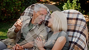 Married senior couple relaxing outdoors in wicker chairs, chatting and kissing