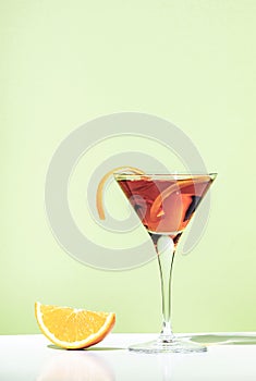 Martinez orange cocktail drink with red vermouth, liqueur, bitter, citrus zest and ice in martini glass. Light green background,