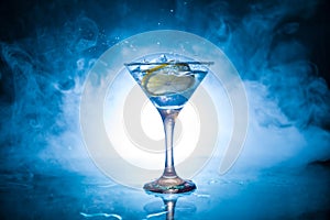 Martini cocktail glass in hand splashing on dark toned smoky background or colorful cocktail in glass with splashes and olives.