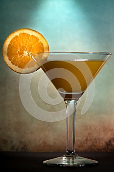 Martini glass with a mix of orange juice and alcohol with a slice of orange fruit against dark and moody orange blue background