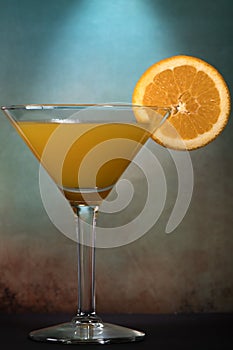 Martini glass with a mix of orange juice and alcohol with a slice of orange fruit against dark and moody orange blue background