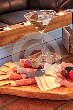 Martini with olives and a Charcuterie board on rustic wood with candles behind a spread of prosciutto panino