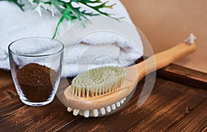 Masage brush, body skin care and coffee natural scrub in glass, white towel and greens on dark wooden background.