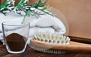 Masage brush, body skin care and coffee natural scrub in glass, white towel and greens on dark wooden background.