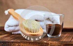 Masage brush, body skin care and coffee natural scrub in glass, white towel and greens on dark wooden background.