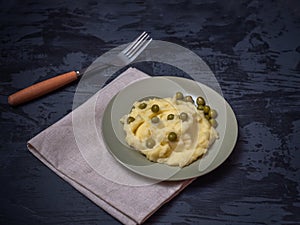 Mashed potatoes with canned peas on a light napkin and a dark wooden background. In the background lies a fork with a wooden spoon