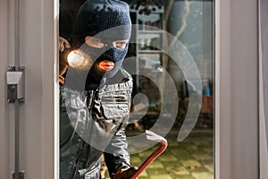 Masked burglar with flashlight and crowbar looking into glass wi