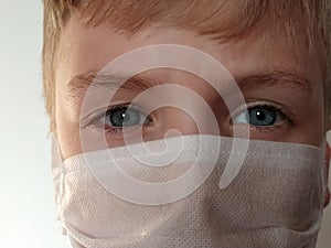 Masked child. The face of a 7-year-old boy in a protective white surgical mask close-up. Schoolboy with blond hair and blue-gray