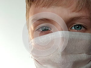 Masked child. The face of a 7-year-old boy in a protective white surgical mask close-up. Schoolboy with blond hair and blue-gray