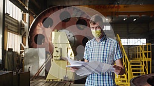Masked engineer examines project plans in industrial workshop. Male professional with blueprint assesses machinery parts