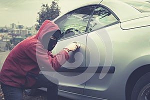 Masked man in casual wear furtively picks the car lock on a city background. Male thief stealthily picklocks the vehicle