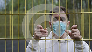 Masked man locked down behind bars in quarantine during pandemic. A man walks up to the fence and looks around