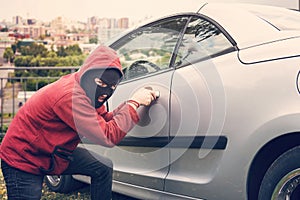 Masked man squats and breaks someone`s car looking to the viewer. Thief in hoodie and hidden face makes attempt to