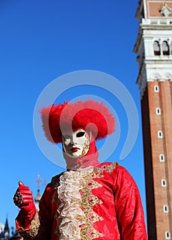 masked person with face covered by mask and red headdress during the Venice carnival with Saint Mark bell tower in the background