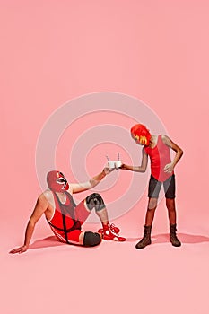 Masked wrestler in red gear seated, toasting milkshakes with standing partner against pink studio background.