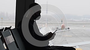 Masked Young Man in Airport Waiting Room Sits and Using a Smartphone by Window