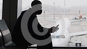 Masked Young Man in Airport Waiting Room Sits and Using a Smartphone by Window