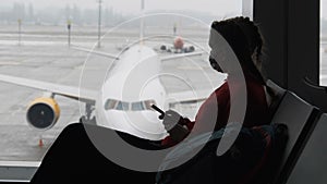 Masked Young Woman in Airport Waiting Room Sits and Using a Smartphone by Window