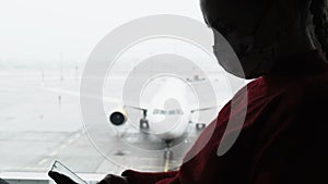 Masked Young Woman in Airport Waiting Room Sits and Using a Smartphone by Window