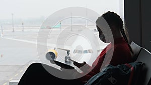 Masked Young Woman in Airport Waiting Room Sits and Using a Smartphone by Window