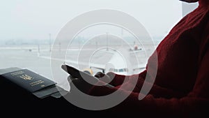 Masked Young Woman in Airport Waiting Room Sits and Using a Smartphone by Window