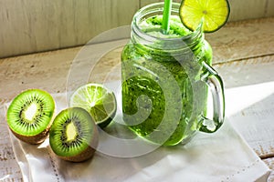 Mason jar mug with green vegetable and fruit smoothie, kiwi, lime, white wood table outdoors, sunlight fleck