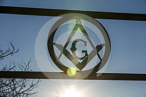 Masonic square and compass sign backlit against a blue sky with sunflare